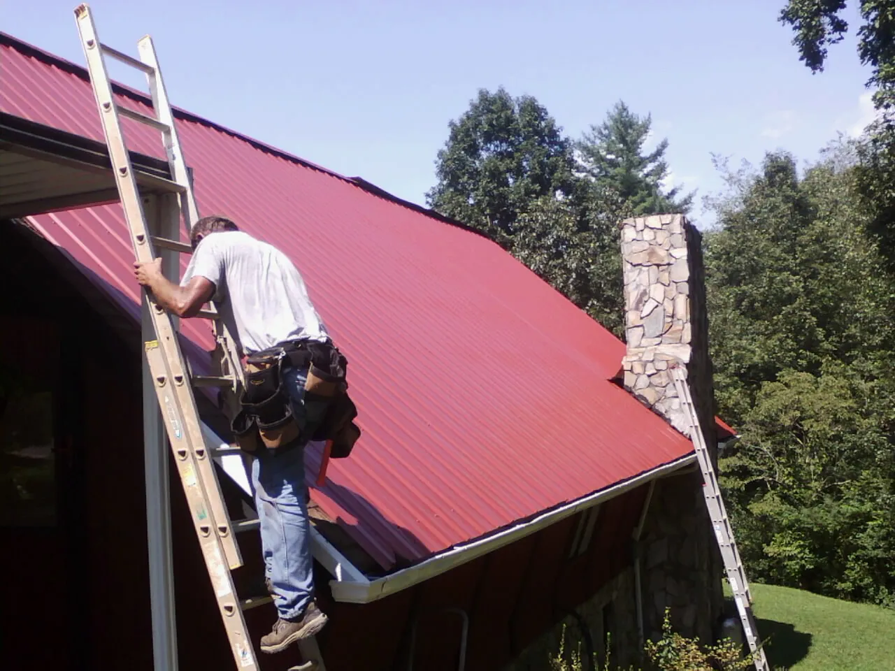 Roofer on a ladder beside a red metal roof and stone chimney — Soffit & Fascia Repair by Roofing Brothers in Harpersville